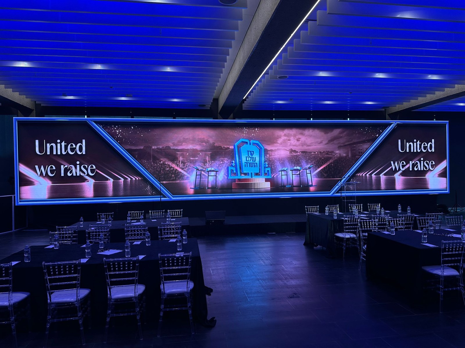 Panoramic LED wall reading United We Raise across a triptych of screens in a blue-lit hall